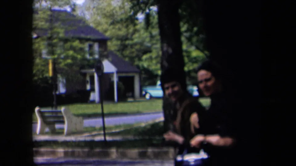 Two adults stand partly in shadow near a tree, with a suburban house, bench, lawn, road, and blue car visible behind them in 1958 New York footage.