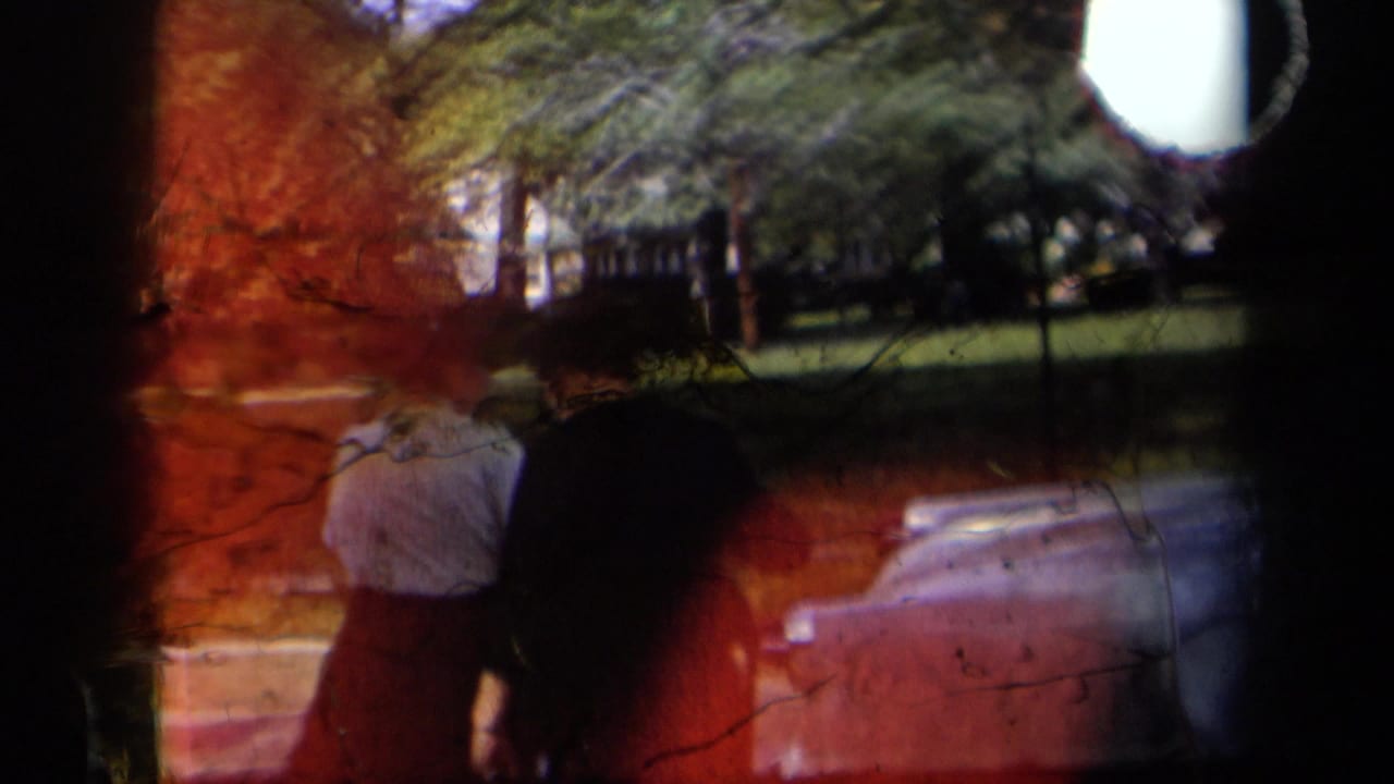 1958 New York home-movie frame showing two men from behind near a parked car in a green park, partly obscured by red film damage.
