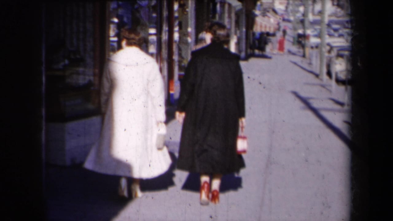 Two women in white and dark coats walk away along a sunny city sidewalk past storefronts.