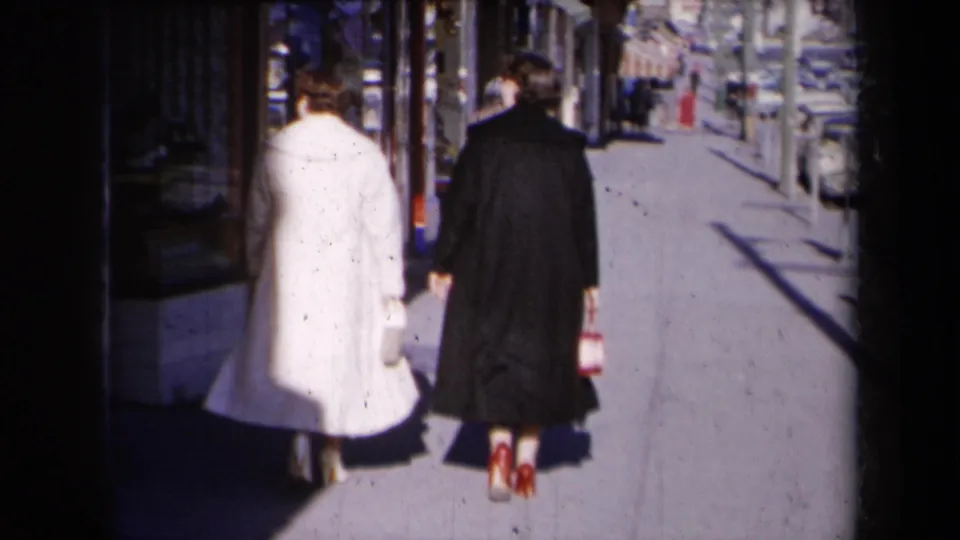 Two women in mid-century coats walk away on a sunlit New York sidewalk, with shop windows and parked cars ahead.