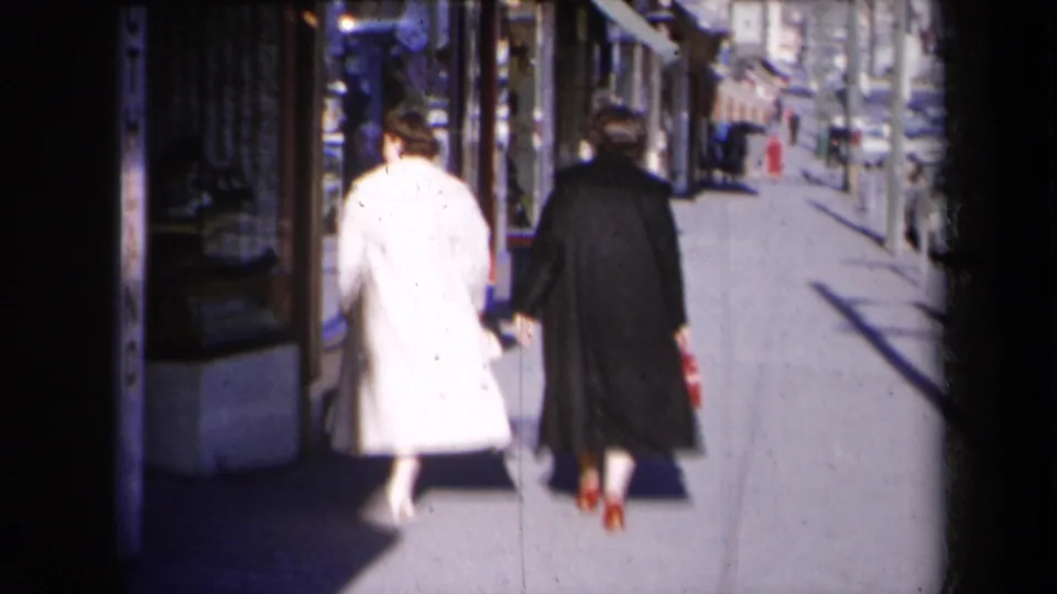 Two women in contrasting coats continue along a New York shopping street, filmed from behind in bright sunlight.