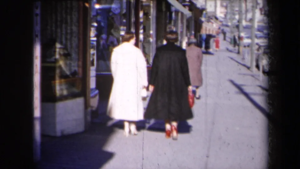 Two women walk past display windows on a 1959 New York sidewalk, with another pedestrian ahead and cars along the street.
