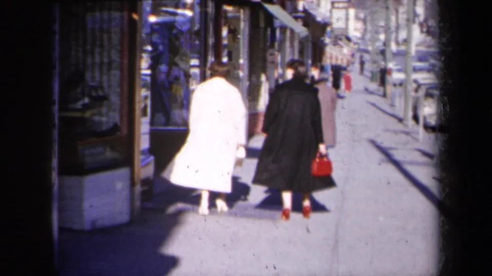 The two women move farther along the New York commercial sidewalk, with a red handbag, shop windows, and parked cars visible.