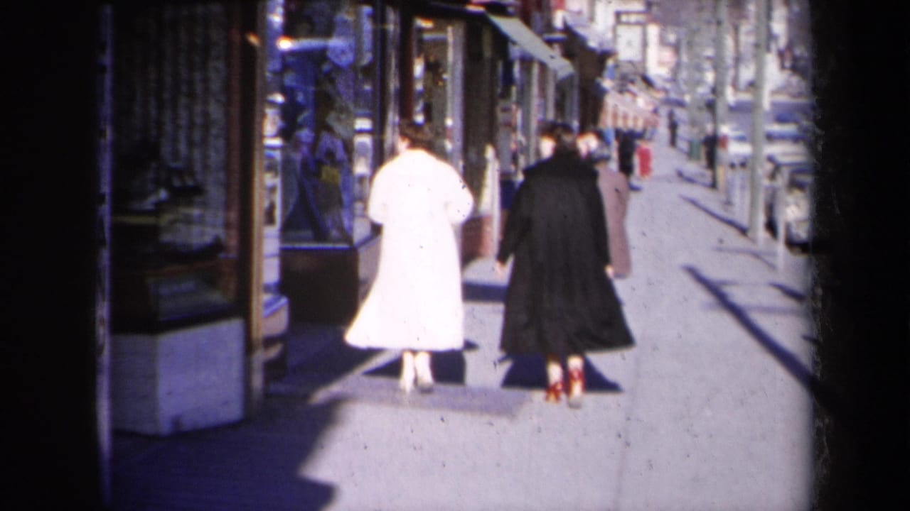 In 1959 New York home-movie footage, two women in white and dark coats walk away from the camera along a bright sidewalk.