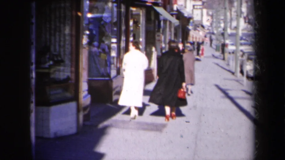 Two women in long coats continue away from the camera on a New York sidewalk with storefronts and curbside cars visible.