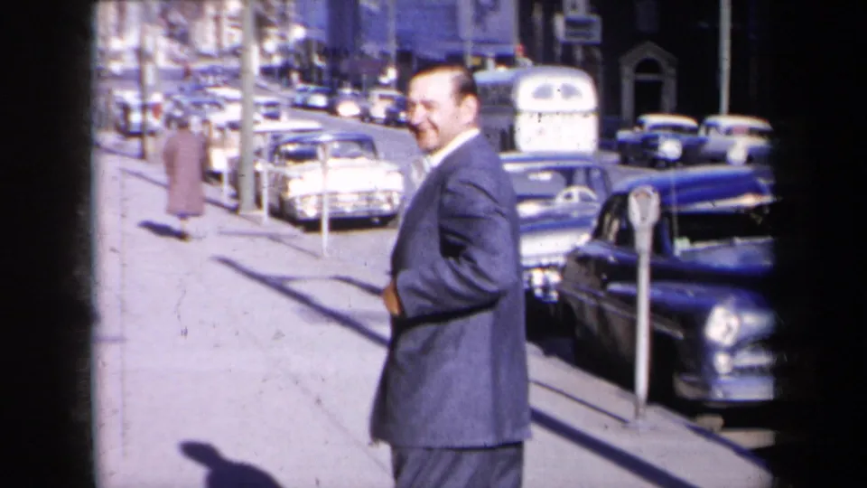 A suited man is seen close to the camera on a 1959 New York sidewalk, with parking meters and late-1950s cars along the curb.
