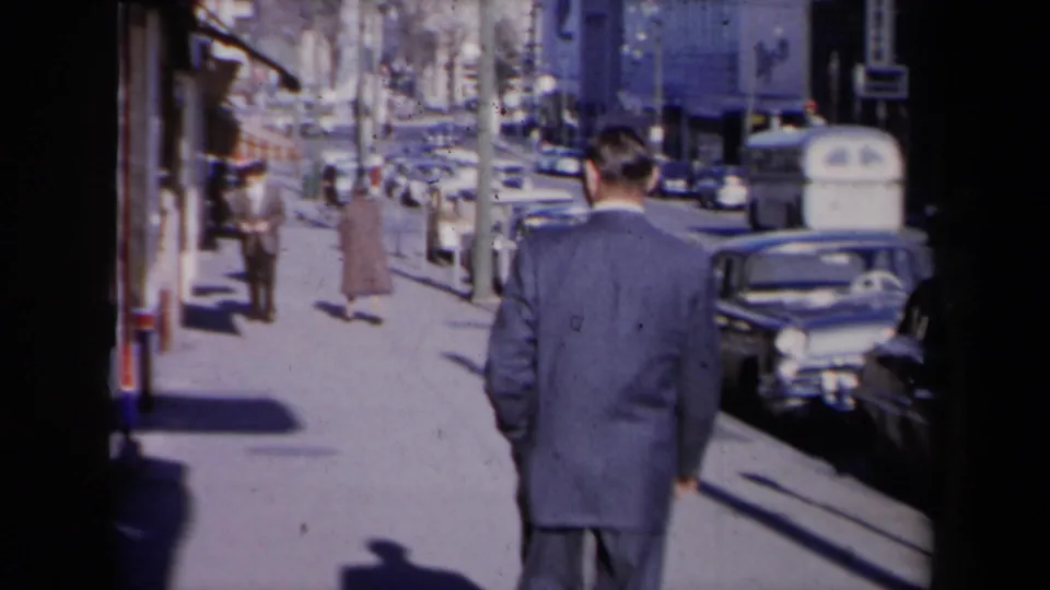 A man in a blue suit walks away on a New York sidewalk while classic cars, parking meters, and pedestrians fill the street scene.