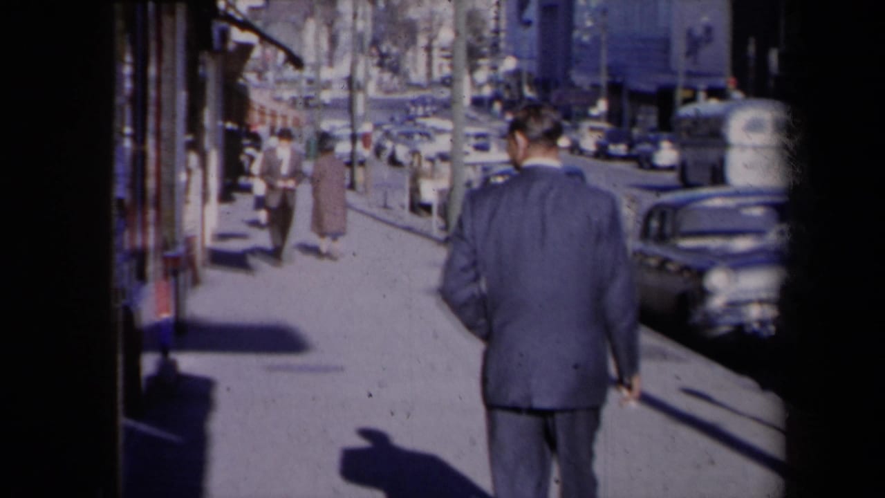 1959 New York street view with a suited man walking away past storefront windows, pedestrians, shadows, and curbside cars.