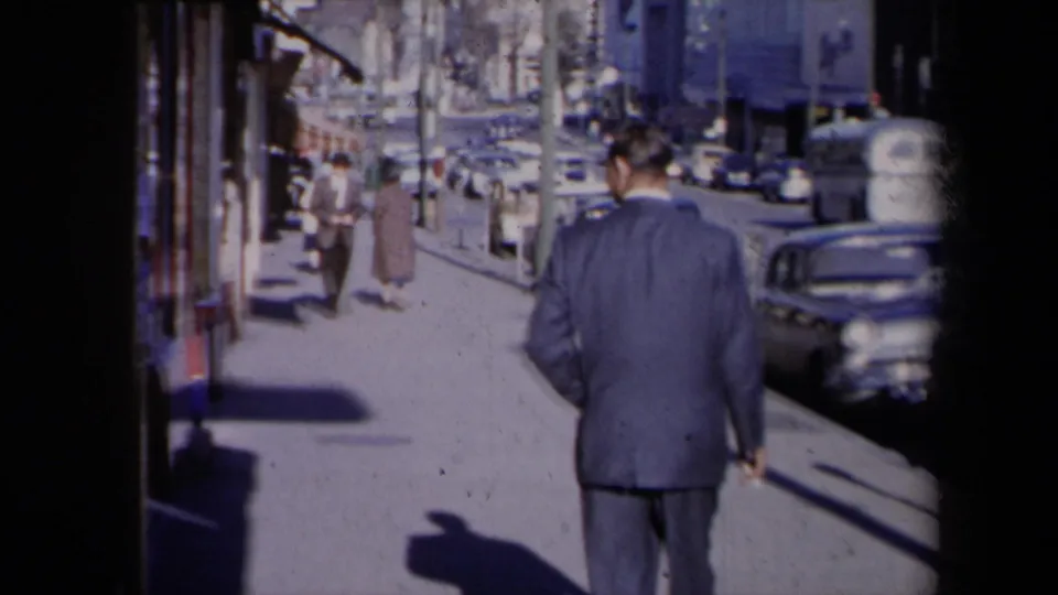1959 New York street view with a suited man walking away past storefront windows, pedestrians, shadows, and curbside cars.