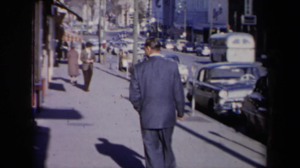 The man in a blue suit walks farther along a New York sidewalk, with parked cars, distant pedestrians, and storefront edges visible.