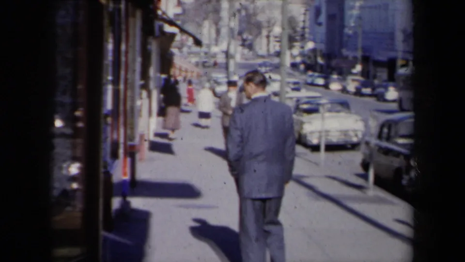 A 1959 New York sidewalk scene shows the suited man from behind, with window reflections, pedestrians, parked cars, and strong shadows.