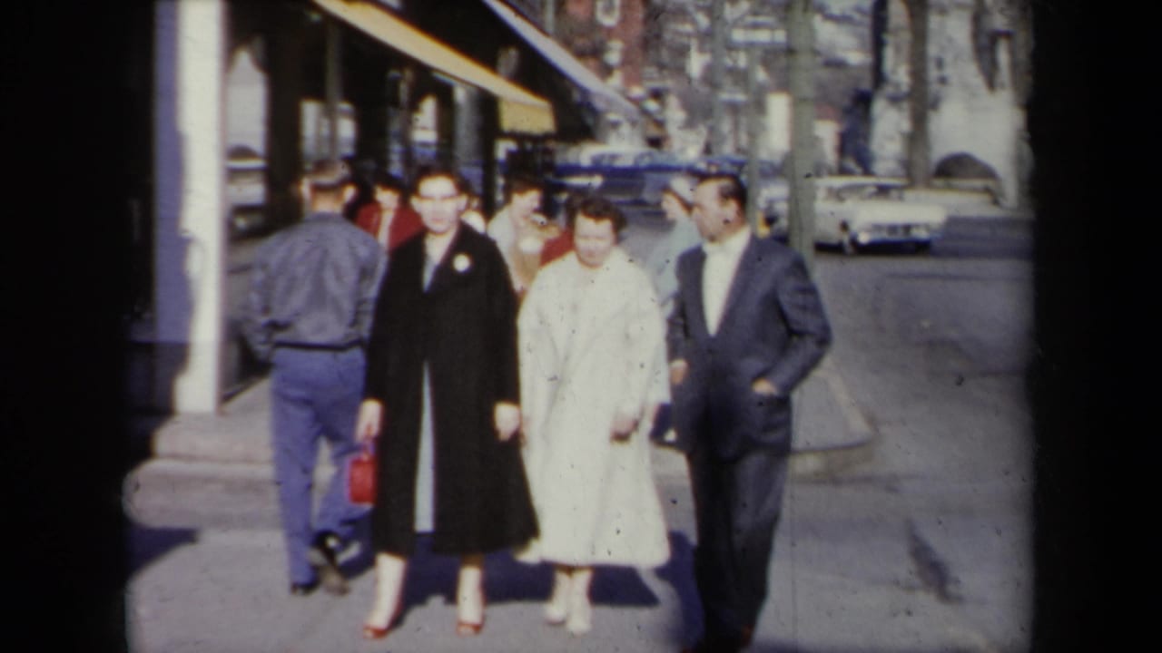 In 1959 New York home-movie footage, two women in coats and a man in a suit walk toward the camera on a sunlit sidewalk.