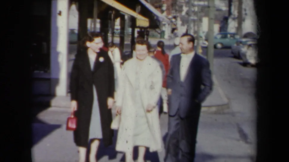 Closer 1959 street view of two women in dress coats and a man in a suit walking on a New York sidewalk.