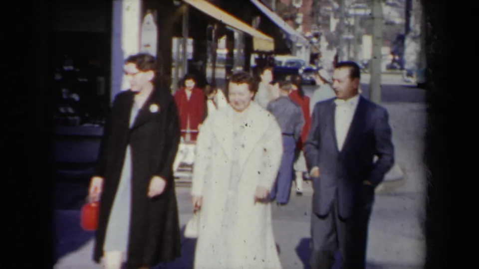 At the end of the short 8mm clip, the adults walk past the storefront while other pedestrians move behind them.