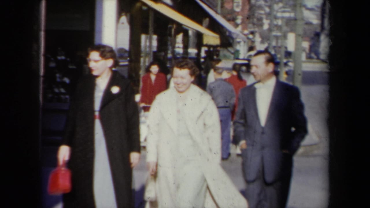 Three well-dressed adults walk along a New York sidewalk in 1959, with storefronts, pedestrians, and parked cars in the background.