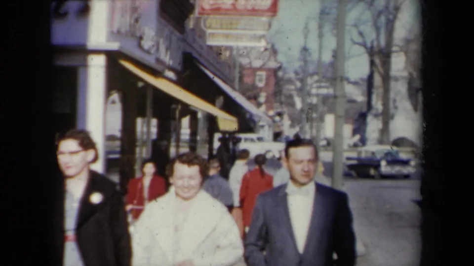 The group walks closer on a 1959 New York sidewalk, with a yellow awning, street signage, pedestrians, and parked cars visible.