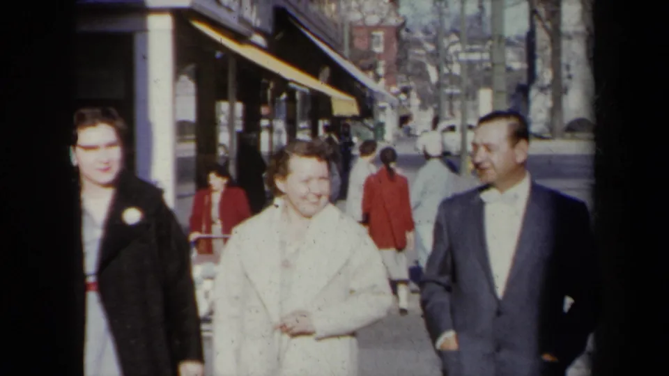 A closer 1959 New York street view shows a woman in a black coat, a smiling woman in a white coat, and a man in a dark suit walking together.