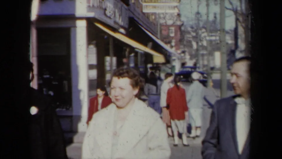 Near the end of the 1959 New York sidewalk shot, the woman in a white coat and the man in a dark suit remain visible with storefronts and pedestrians behind.