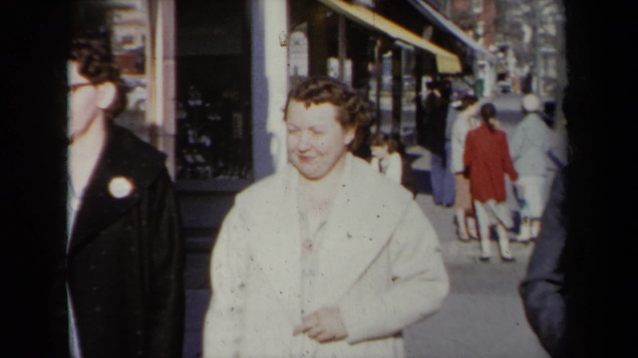 1959 New York street sidewalk view with pedestrians in coats passing storefront windows in soft 8mm home-movie color.