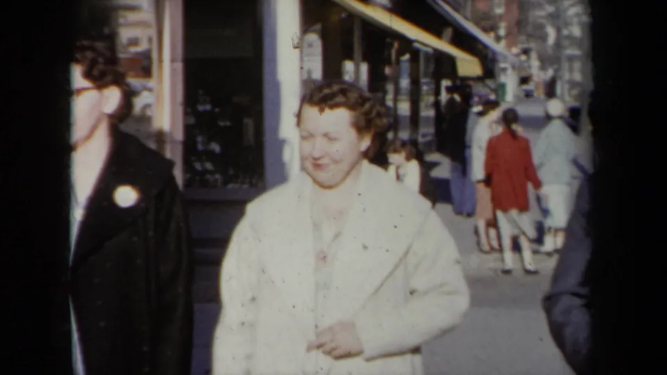 1959 New York street sidewalk view with pedestrians in coats passing storefront windows in soft 8mm home-movie color.
