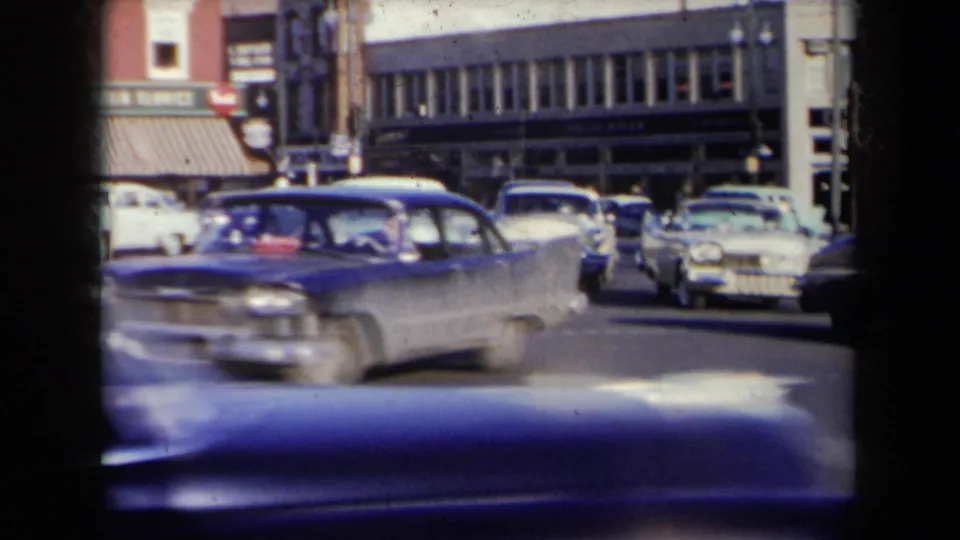 Late-1950s cars pass through a New York intersection, including a blue sedan and lighter cars beyond it.