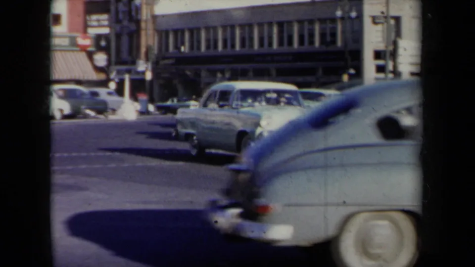 A rounded mid-century car crosses the foreground as additional late-1950s vehicles approach on a New York street.