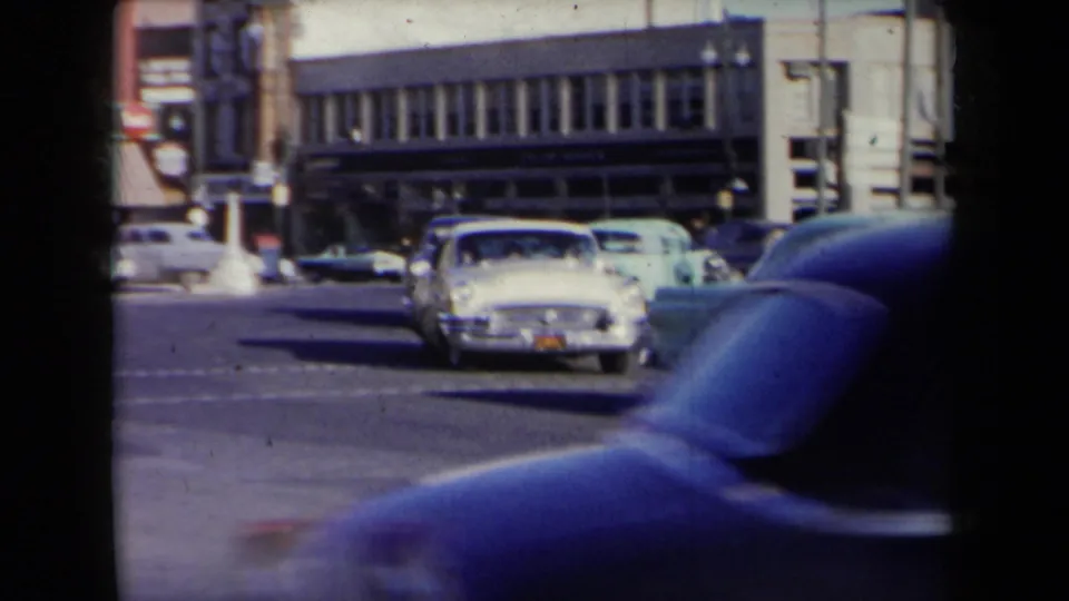 A white convertible and other 1950s cars move through a New York intersection, partly obscured by a blue foreground car.