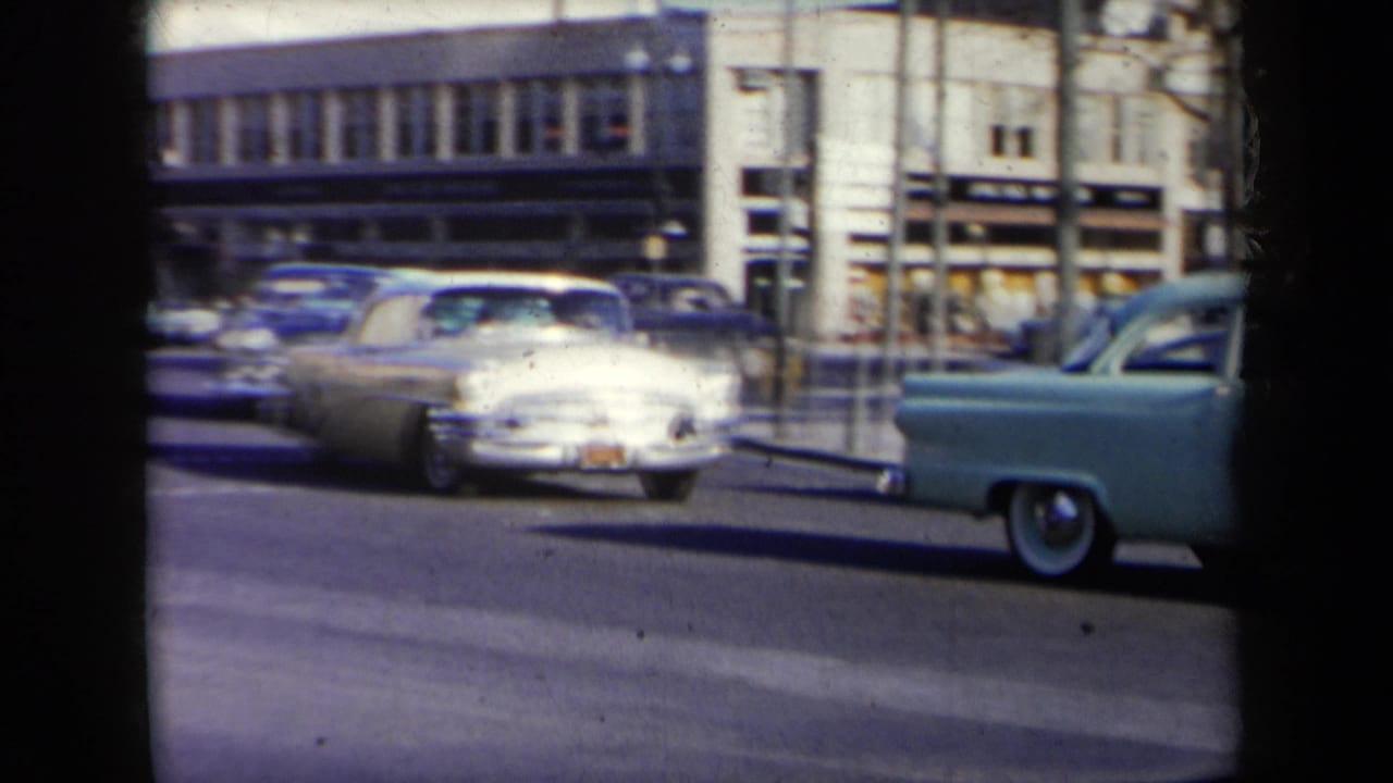 Classic cars pass along a busy city street with storefronts in the background.