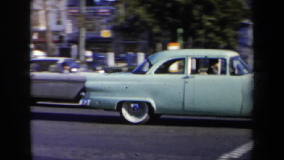 Light blue mid-century sedan crosses a New York city street, with traffic and buildings visible behind it.