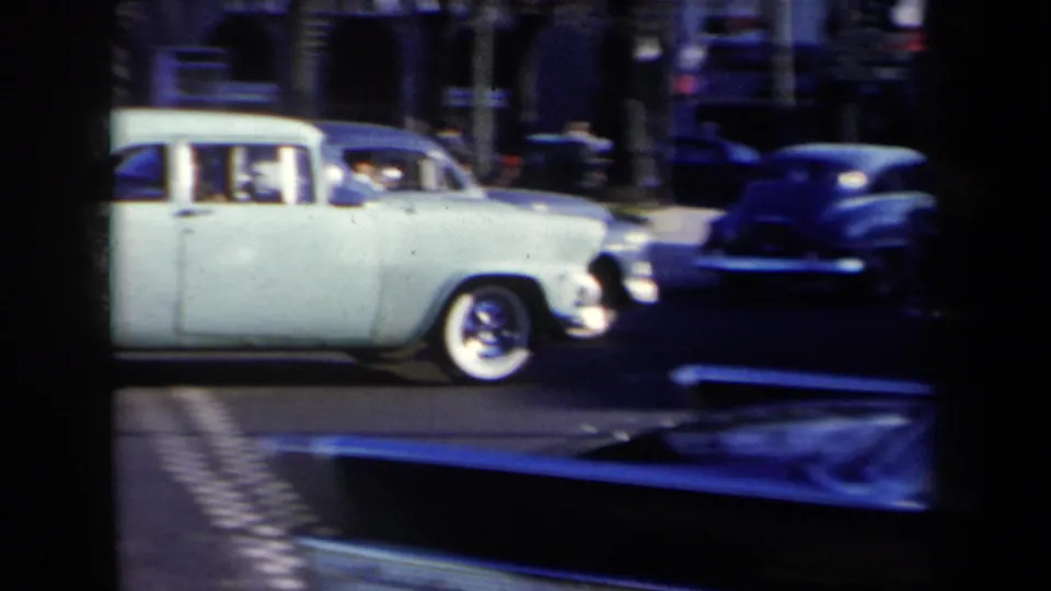 A pale 1950s sedan moves through a New York intersection while darker cars pass behind it.