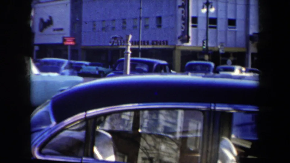 Foreground view of a dark mid-century car with New York traffic and building facades beyond it.
