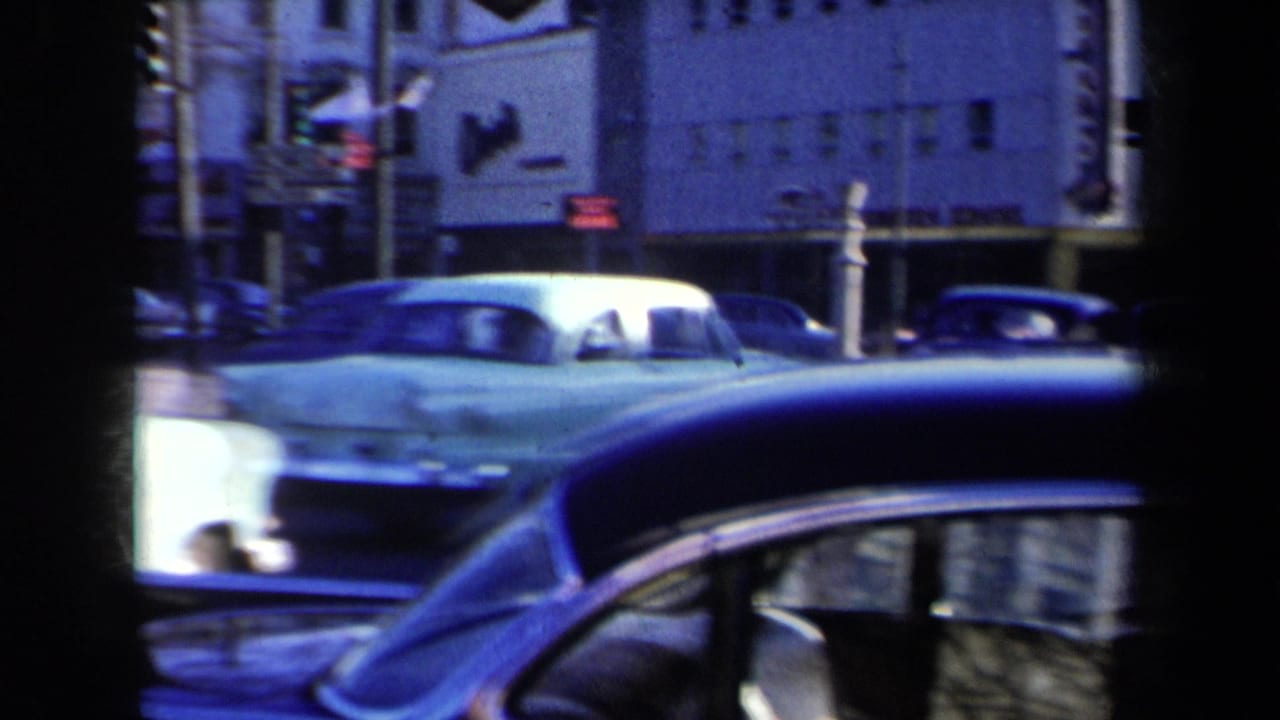1959 New York street view with late-1950s cars moving past storefronts, traffic lights, and the edge of another car in the foreground.