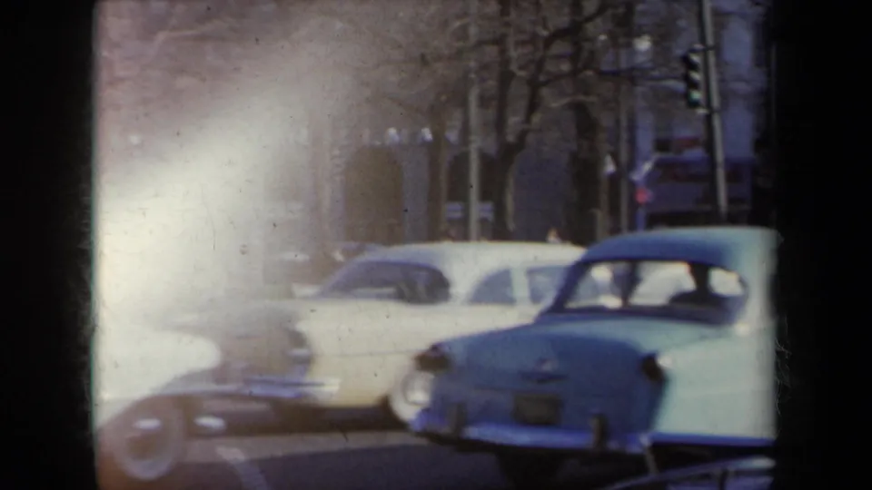 1959 New York traffic scene with pale sedans and a station wagon moving past bare trees and shadowed building fronts.