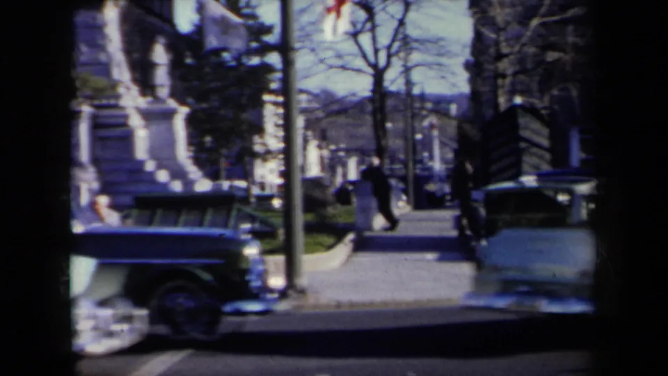 1959 New York sidewalk view with pedestrians near a large stone monument while cars pass in the foreground.