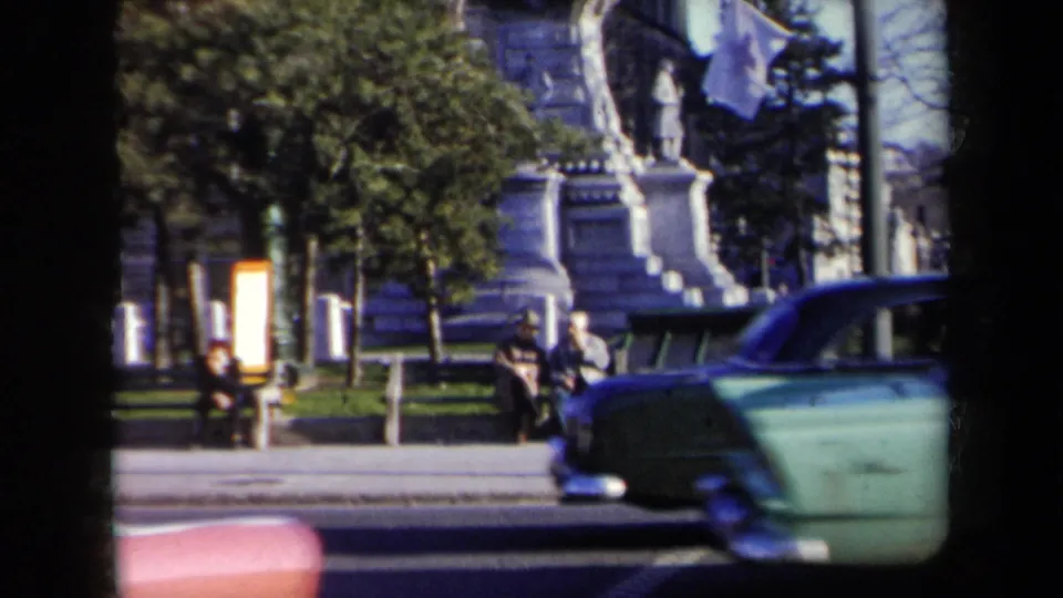 1959 New York street frame with passing cars, seated pedestrians, trees, benches, and part of a large stone monument.