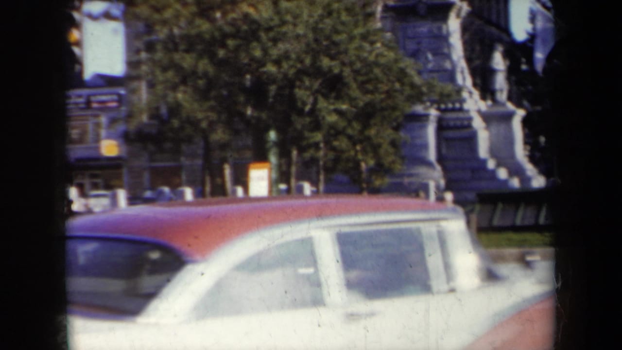 Red-and-white sedan passing a downtown intersection with a monument and storefronts behind it.