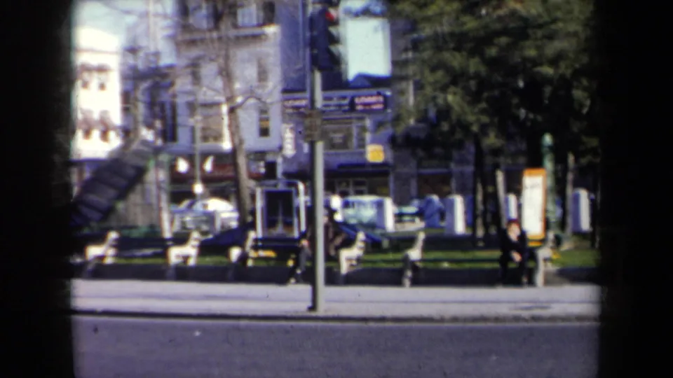 1959 New York street view with a monument base, parked cars, pedestrians, trees, and commercial buildings in the background.