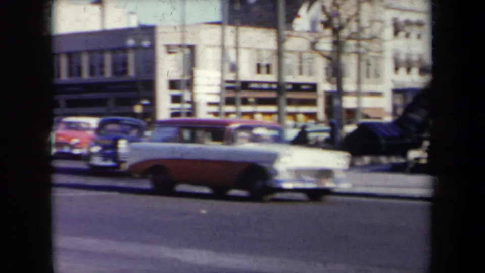 1959 New York frame showing a two-tone station wagon and other mid-century cars passing commercial storefronts.