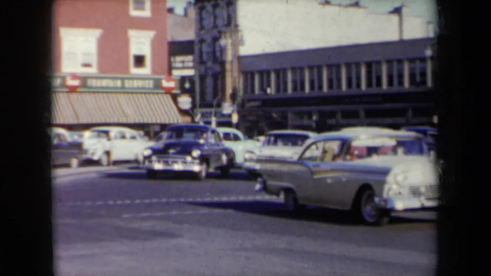 1959 New York downtown intersection with multiple period cars, visible shop awnings, street poles, and commercial buildings.