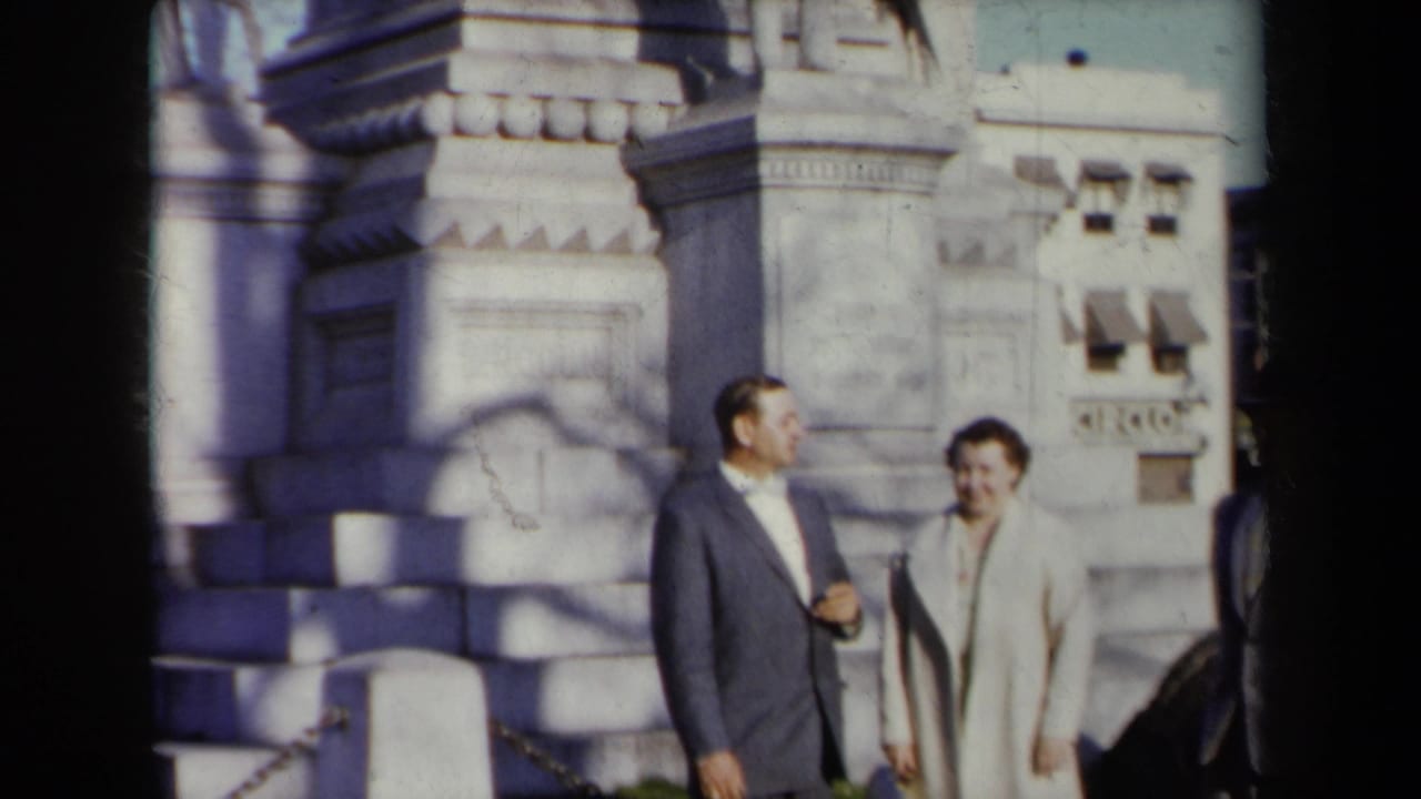 In 1959 New York home-movie footage, a suited man and a woman in a light coat stand before a carved stone monument.