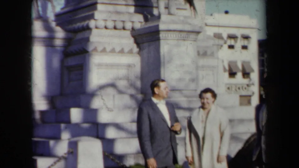 In 1959 New York home-movie footage, a suited man and a woman in a light coat stand before a carved stone monument.