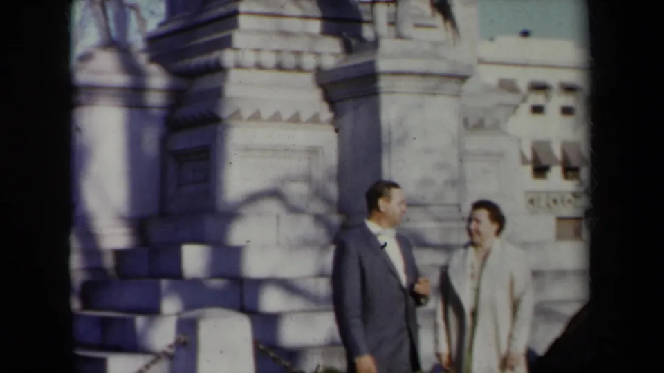 The suited man and woman remain beside the stone monument, with soft 8mm color and visible architectural carving.