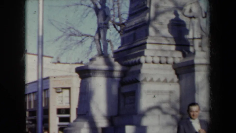 The handheld camera moves up the monument, showing stone columns, bare branches, nearby buildings, and part of a suited man at the bottom edge.