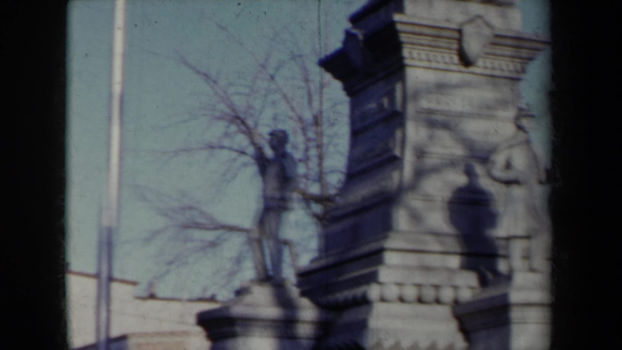 1959 New York home-movie frame showing a stone monument with a soldier figure, pedestal masonry, bare trees, and nearby buildings.