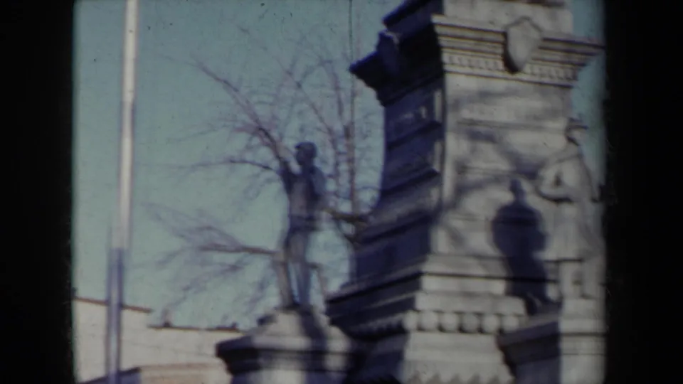 1959 New York home-movie frame showing a stone monument with a soldier figure, pedestal masonry, bare trees, and nearby buildings.