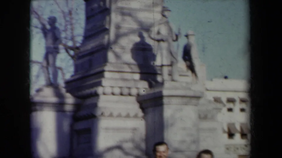 1959 New York frame of a stone monument with sculpted figures and pedestal blocks, with two adults partly visible near the bottom edge.