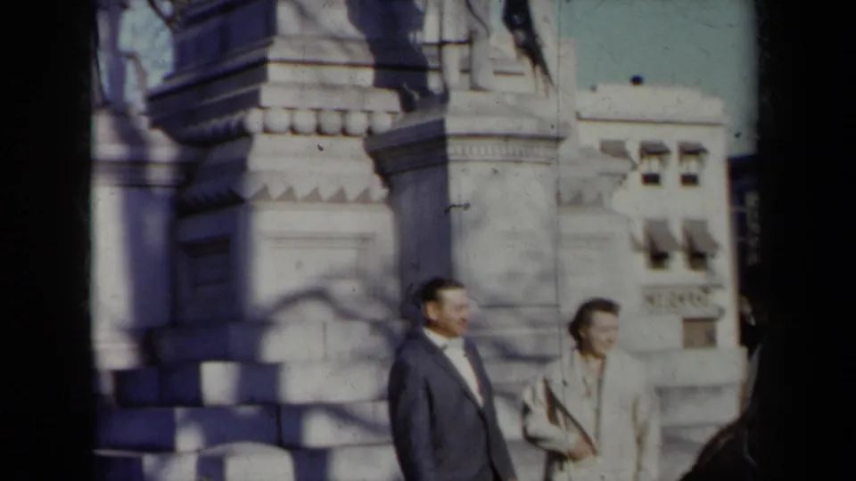 1959 New York home-movie frame showing a suited man and a woman in a light coat standing by a large stone monument.