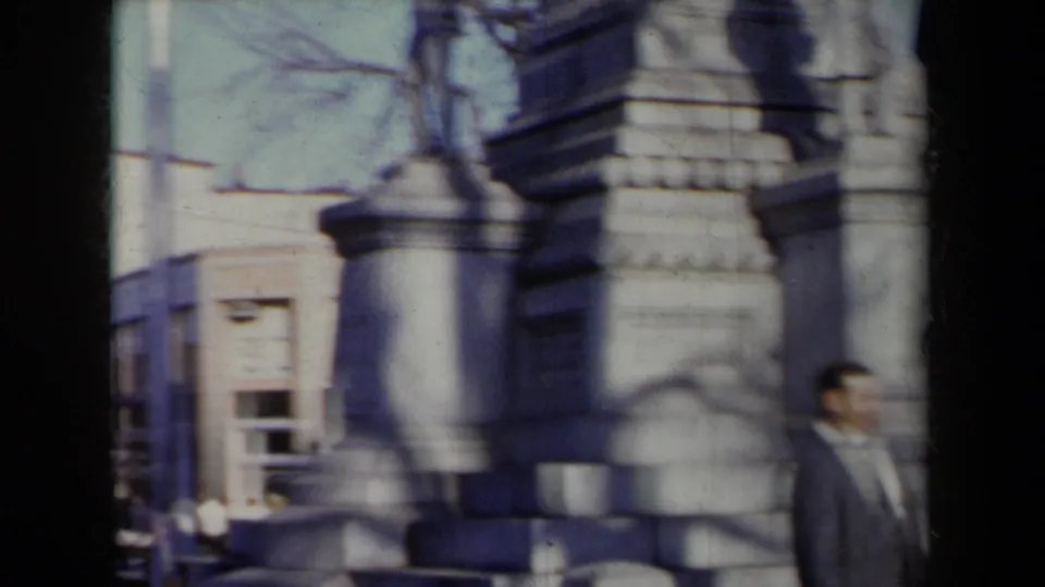 1959 New York frame showing a stone monument, soldier figure, pedestal masonry, bare trees, and a suited man at the edge.