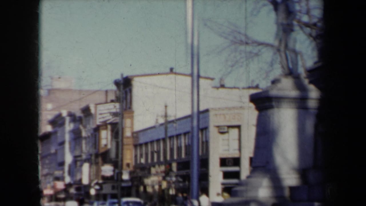 1959 New York street view with commercial buildings, bare winter trees, cars, pedestrians, and a stone monument partly crossing the frame.