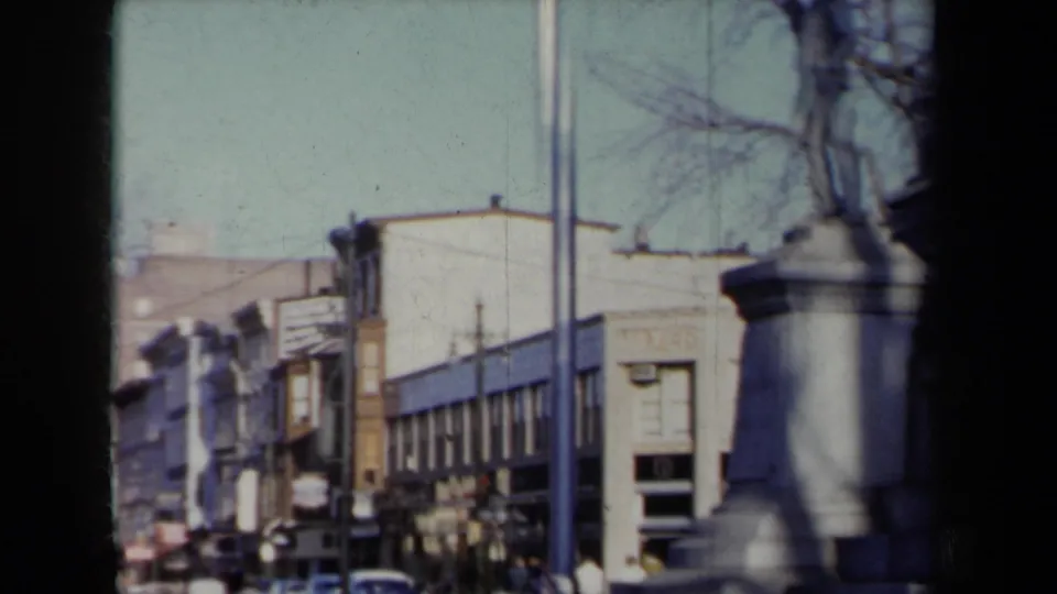 1959 New York street view with commercial buildings, bare winter trees, cars, pedestrians, and a stone monument partly crossing the frame.